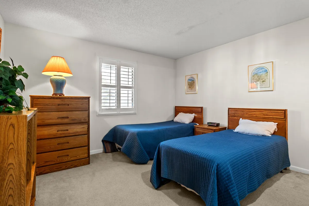 A charming second bedroom with two twin beds dressed in blue covers, a wooden dresser, a warm table lamp, and framed artwork on the walls. The room feels bright and cozy, ideal for family members or friends seeking privacy and comfort.