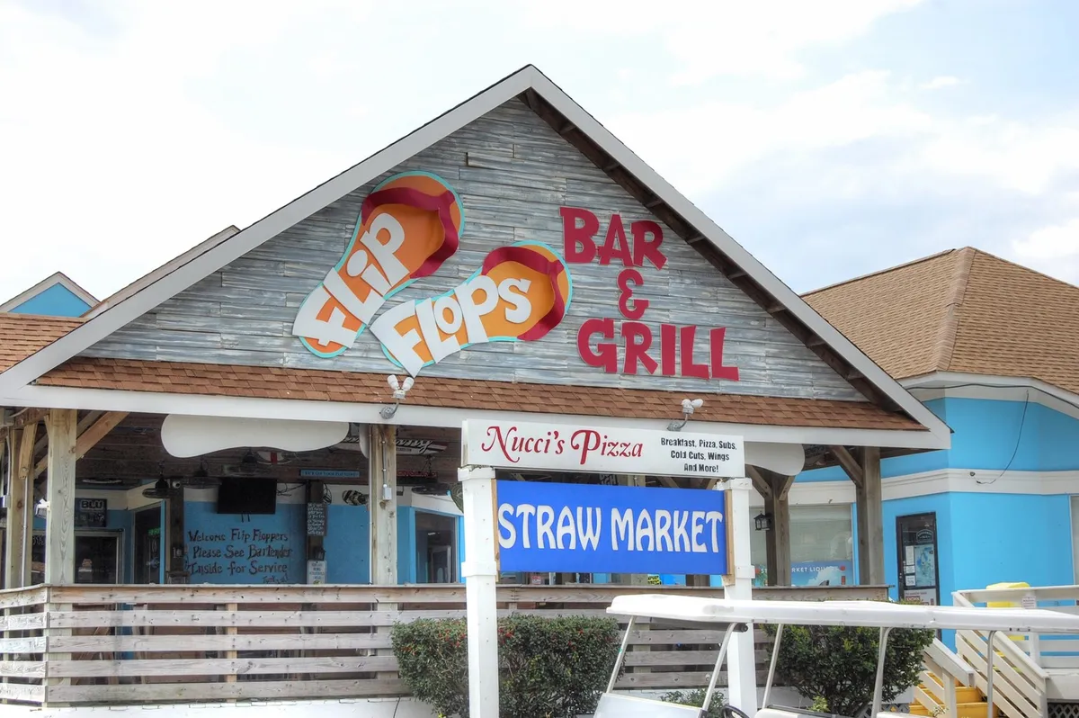 A vibrant shot of the local Straw Market area, featuring the Flip Flops Bar & Grill, Nucci's Pizza, and Straw Market signage. The colorful facades and sunny atmosphere evoke fun, social evenings out, with opportunities for dining, drinks, and mingling just minutes away from the retreat.