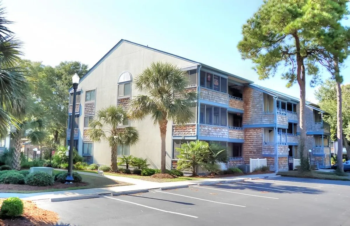 The image features an inviting exterior view of the property, with palm trees framing a multi-story building adorned with balconies. Stone facades, a landscaped setting, and a visible parking area create a welcoming, resort-like atmosphere that hints at private balconies for morning coffee or evening relaxation.