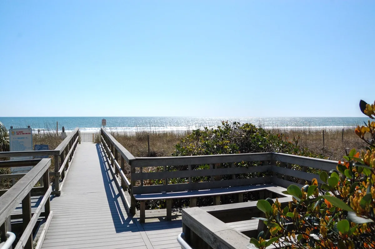A serene wooden walkway leads through dune grass and coastal shrubs toward the open ocean, inviting families to stroll from their condo to the beach. The scene hints at the easy, relaxing transition from accommodation to the natural beauty of the shore.