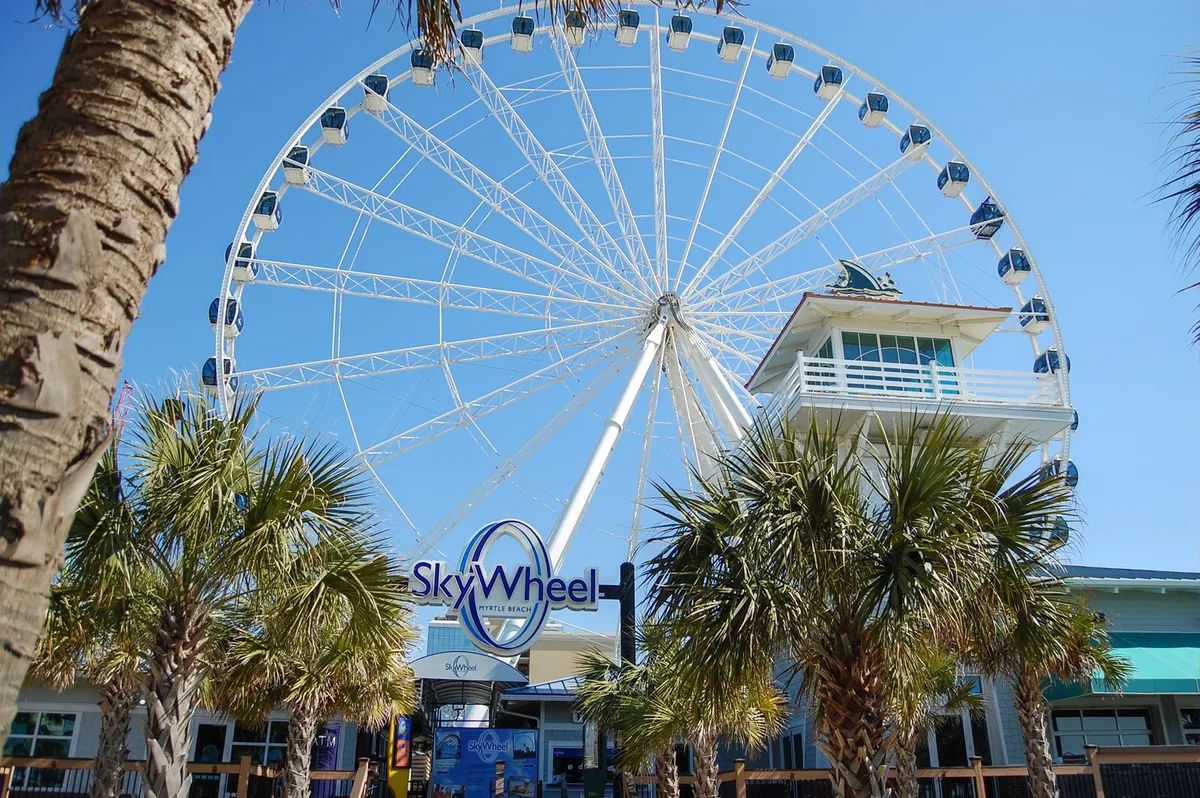 The iconic Myrtle Beach SkyWheel rises above palm trees with its ticket booth in the foreground, set against a bright sky. This lively scene captures one of the area’s top attractions and highlights the fun activities available through the Free Fun Program.