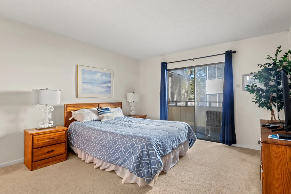 A tranquil bedroom featuring a cozy Queen bed, a large window letting in natural light, soft linens, nightstands with lamps, and tasteful wall art. The serene color palette and inviting decor create a restful atmosphere, perfect for winding down after a day at the beach.