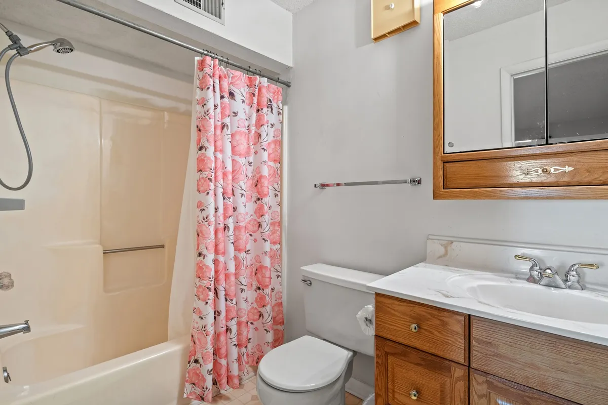 A modern, inviting bathroom with a flower-patterned shower curtain, wooden vanity, well-lit mirror, and practical storage. The space feels fresh and welcoming—ideal for morning routines or winding down with an evening bath.