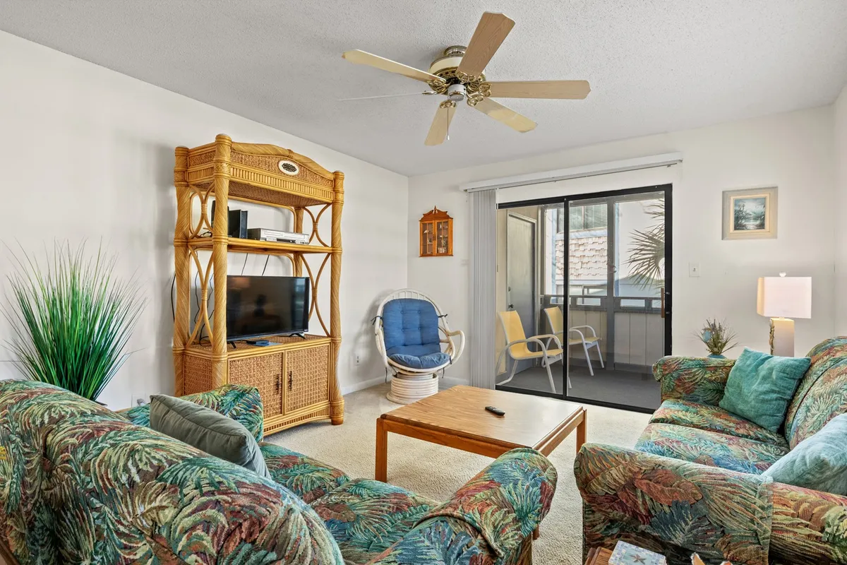 A spacious living room with tropical-patterned sofas, rattan furniture, a central coffee table, and a television. Sunlight streams in, creating a bright and comfortable setting. The furniture is arranged to encourage conversation and relaxation, perfect for group gatherings or watching a movie together.