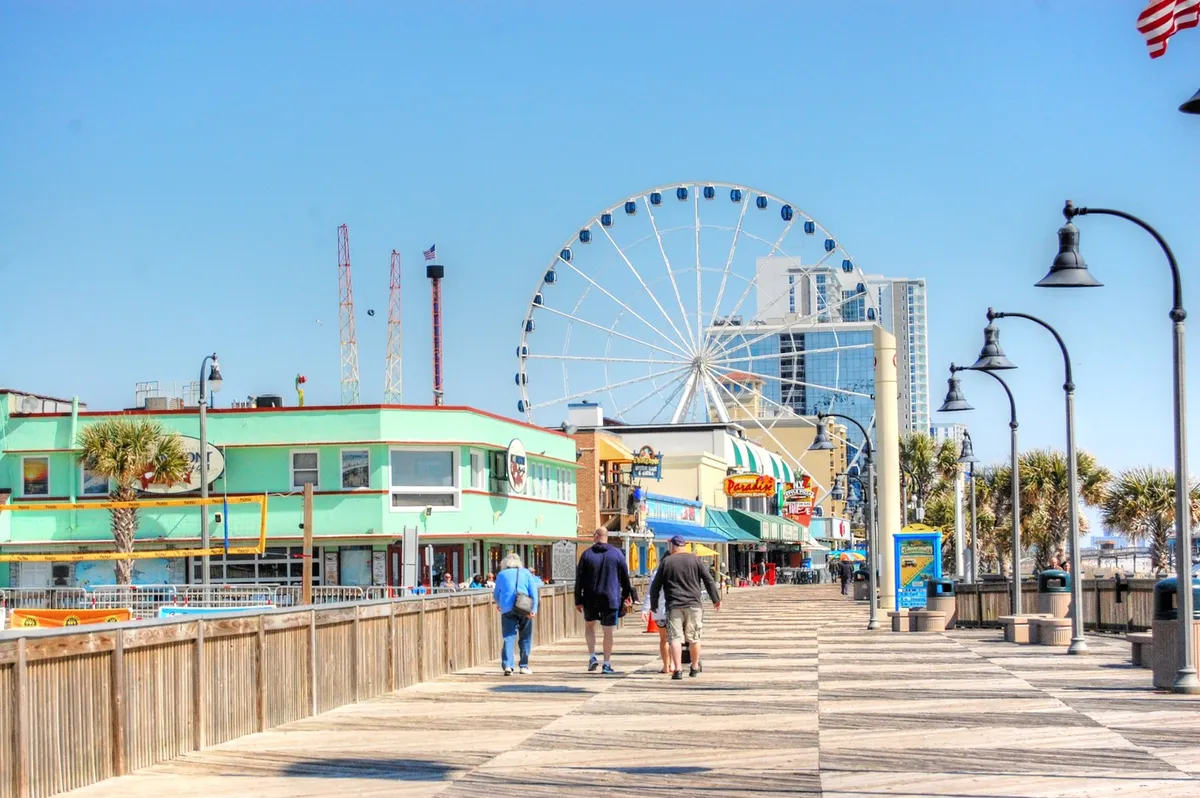 This lively exterior image captures a ferris wheel, colorful boardwalk buildings, and people strolling under a bright sky, evoking the excitement and variety of group outings in the area. The boardwalk setting reflects Myrtle Beach’s signature attractions, including shopping, entertainment, and family-friendly adventure.