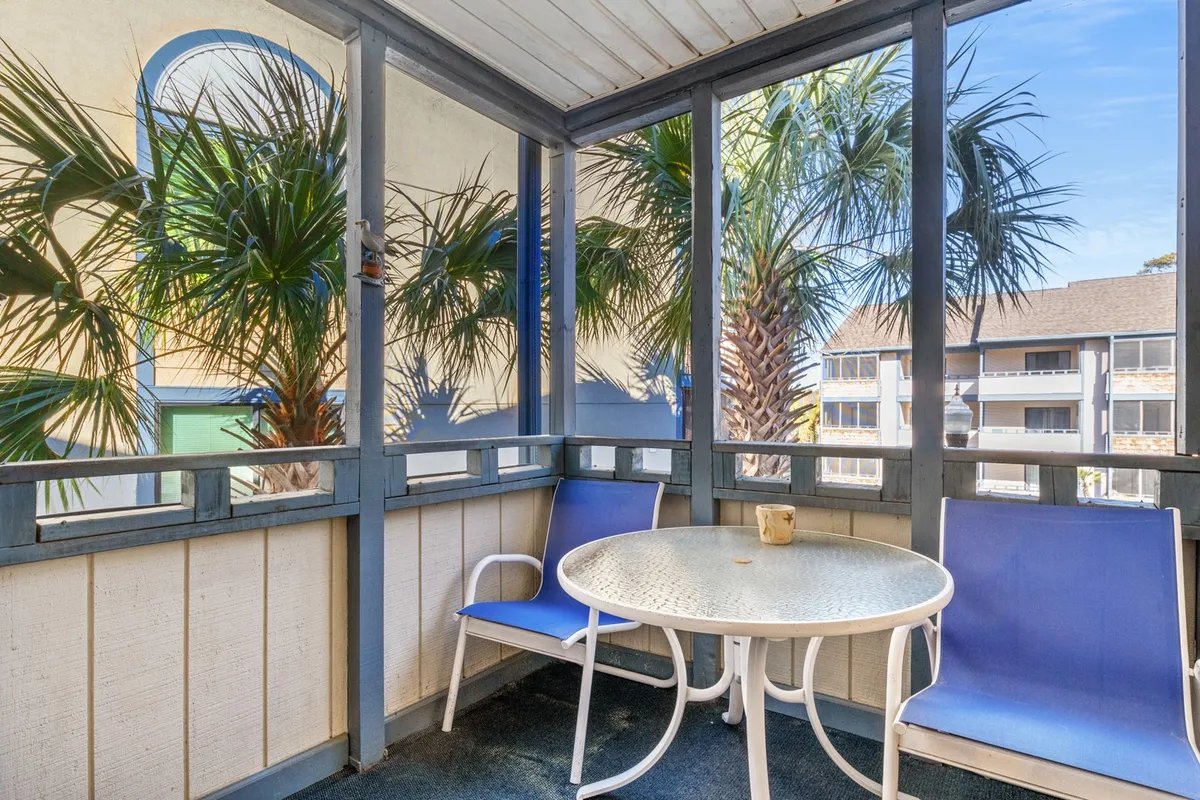 A tranquil screened-in porch featuring a table and chairs, surrounded by a screened enclosure with palm trees visible outside. The scene is set for enjoying morning coffee, reading a book, or relaxing with friends, capturing the breezy indoor-outdoor comfort of the space.