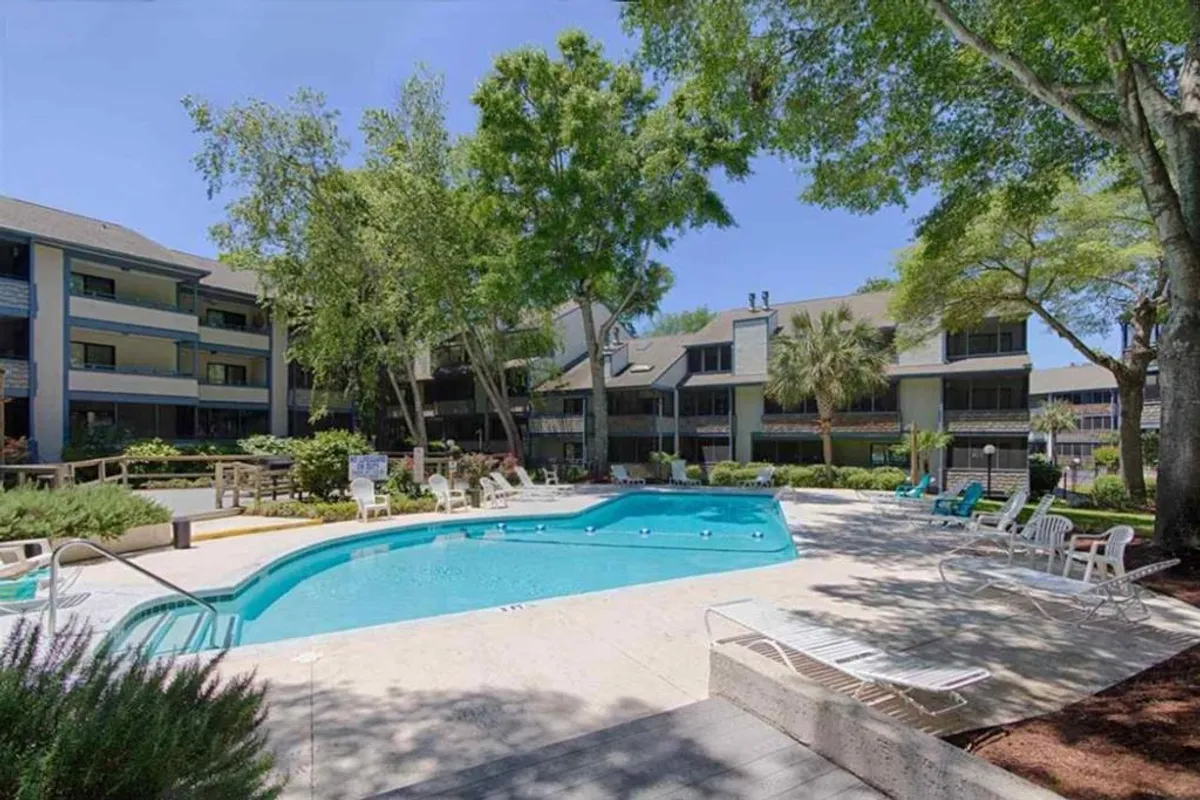 A sparkling outdoor swimming pool surrounded by lounge chairs and lush greenery, set against the backdrop of the building exterior. The area suggests opportunities for sunbathing, swimming, and social gatherings in a leisurely resort atmosphere.