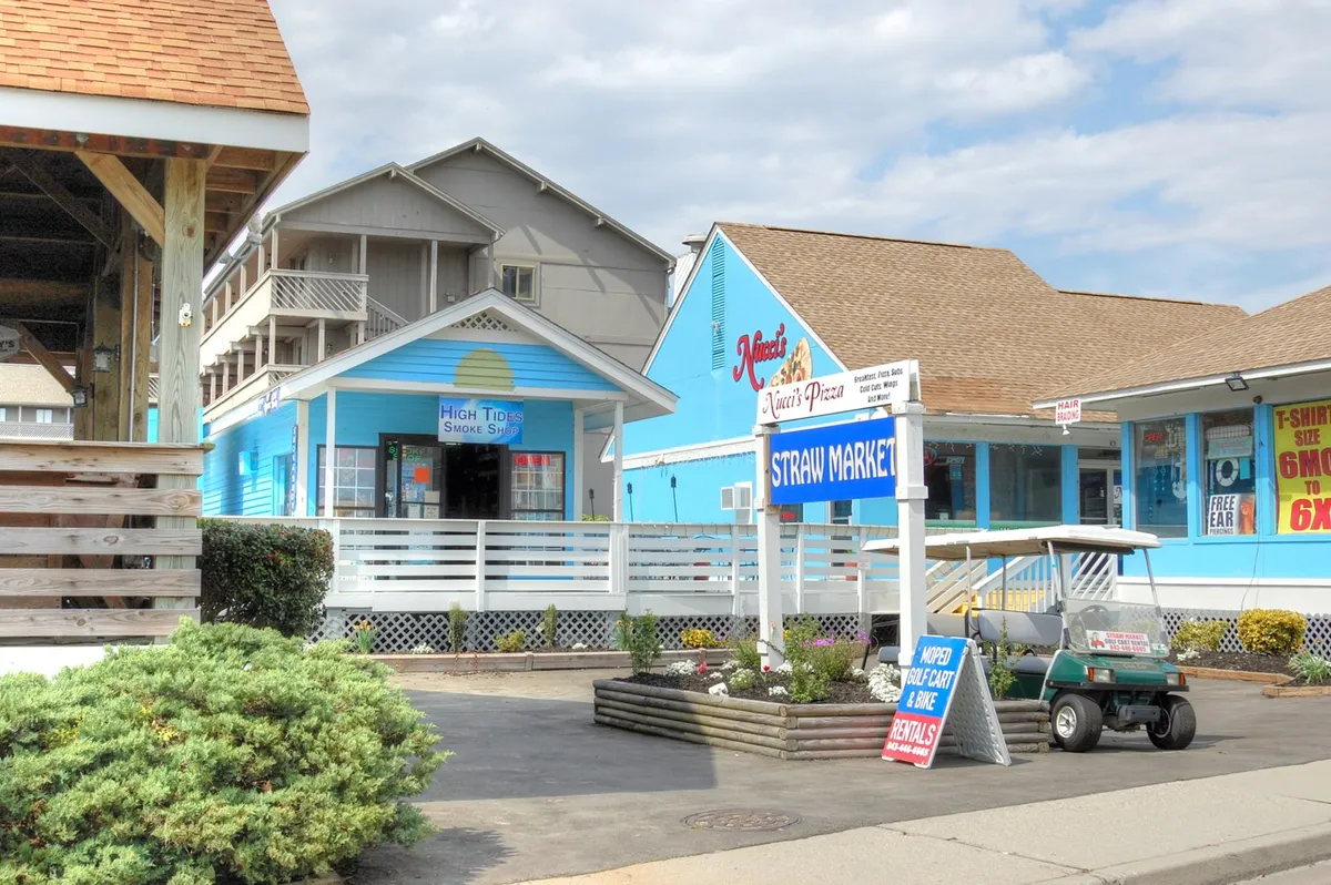 A bustling exterior scene featuring colorful market facades, the ‘Straw Market’ sign, and nearby local eateries like Nino’s Pizza and High Tide Smoke Shop. The lively atmosphere suggests opportunities for shopping, dining, and exploring community events.